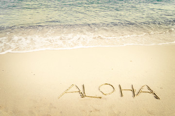 Inscription Aloha written on the sandy beach with ocean wave - free space. vintage color tone effect