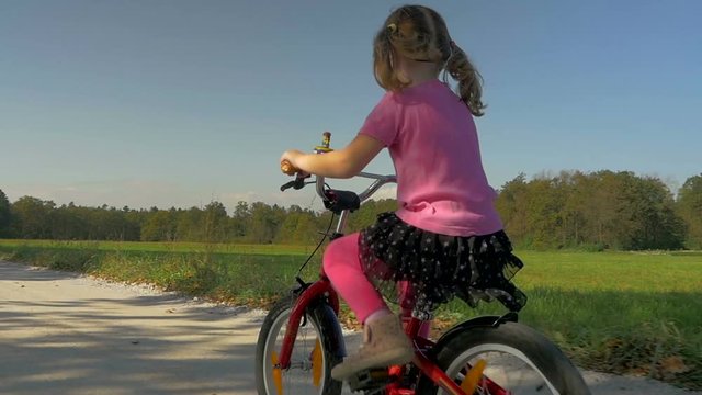 Low angle, dolly shot of a cute little girl how rides her red bike on the macadam road
