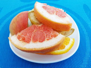 Slices of grapefruit, orange, banana and apple on a white plate, which stands on a blue tablecloth