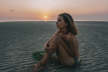 Fototapeta na wymiar Young pretty girl sitting on the sand during sunset with palm branch