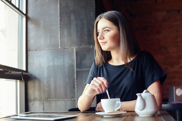 Portrait of young cute girl stirring sugar in hot tea and thoughtfully looking out of the coffee...
