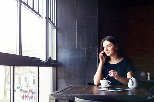 Young attractive girl talking on mobile phone and smilling while sitting alone in coffee shop during free time. happy female having rest in cafe. Lifestyle, coffee