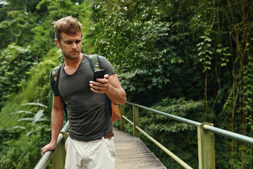Naklejka na ściany i meble Man Using Mobile Phone For Communication. Tourist Male Holding Smartphone On Summer Travel Vacation. Beautiful Handsome Happy Traveler Enjoying Tropical Forest Nature. Tourism, Technology Connection. 