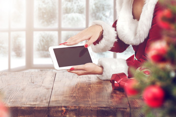 Female Santa's helper checking gift list on tablet at wooden desk, Christmas holiday concept. Snowy...