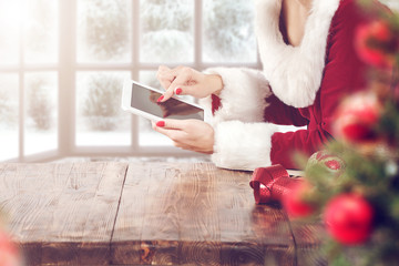 Female Santa's helper checking gift list on tablet at wooden desk, Christmas holiday concept. Snowy...