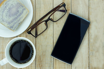 Smartphone, eyeglasses and a cup of coffee with bread on wooden