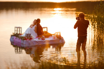 Wedding photographer in action, taking a picture of the bride and groom sitting on the raft. Summer, sunset - obrazy, fototapety, plakaty