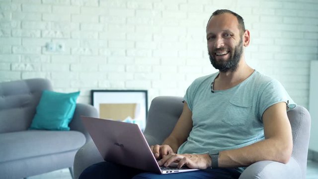 Happy man with laptop talking to camera while sitting in armchair at home
