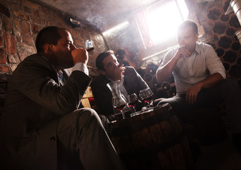 Three businessmen sitting in the wine cellar drinking wine and smoke cigar, resting after a hard day at work.