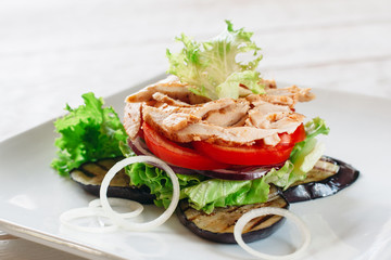 Layered salad with chicken, tomato and grilled eggplant on white. White square plate with portion of layered salad with fried chicken strips, tomato, lettuce, onion and grilled eggplant.