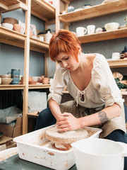 Woman works on potters wheel at studio. Plenty of pottery on background. Young red beautiful woman intently work on potters wheeln potters wheel