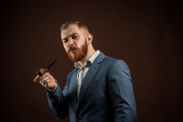 Portrait of handsome confident man in suit holding smoking pipe against of brown...