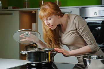 Young woman in the kitchen. Beautiful young housewife looking in to pan. Red-haired girl smiles...