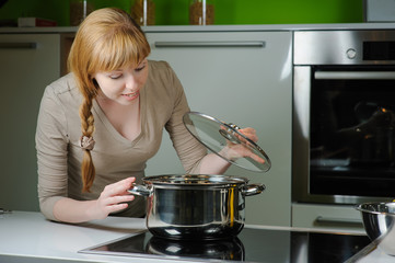Young woman in the kitchen. Beautiful young housewife looking in to pan. Red-haired girl smiles...