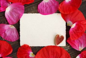 Empty white ancient photograph with decorative heart on a wooden surface in the scattered pink poppy petals. Romantic background by St. Valentine's Day, a wedding.
