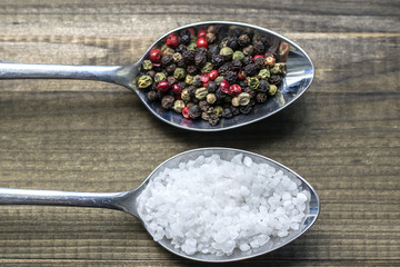 Salt and pepper. Two metal spoons with salt crystals and color peppercorns on wooden table