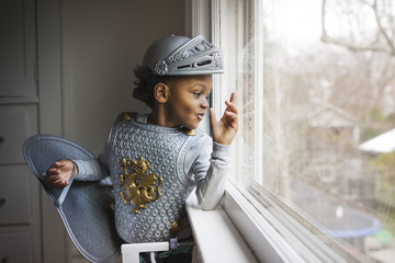 Little boy dressed up in armor costume looking out through window at home