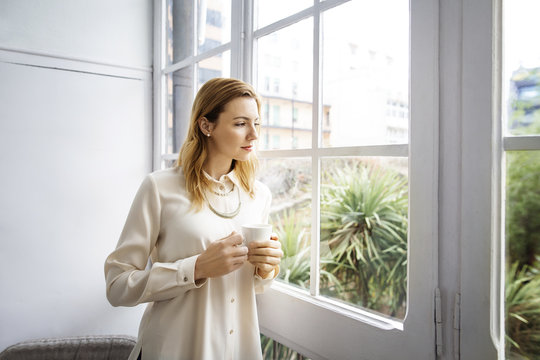 Businesswoman looking through window while holding coffee cup in office