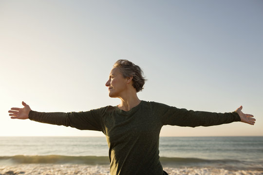 Senior woman with arms outstretched standing on beach against clear sky