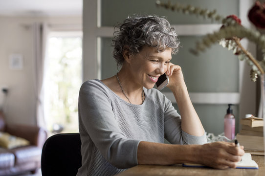 Mature woman talking on smart phone and writing in book at home