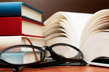 Close up of stack of books, large book pages and glasses.
Glasses and open book on wooden table.
