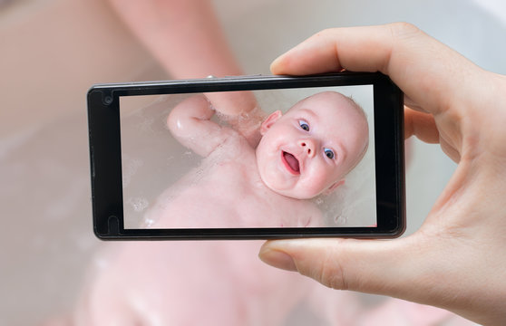 Parent is taking photo of a happy baby in bath with smartphone.