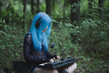 Young girl with blue hair playing synthesizer dressed in black sitting on grass in spring in park with copy space. Female composer and musician. Creative woman
