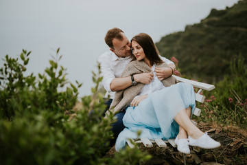 Young cute couple honeymoon on a white chair holding their hands on dating in a beautiful place italy near ocean and mountains, hug, smile and talk to each other
