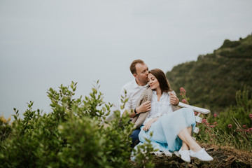 Young cute couple honeymoon on a white chair holding their hands on dating in a beautiful place italy near ocean and mountains, hug, smile and talk to each other
