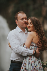 Young cute couple honeymoon standing near waterfall holding their hands on dating in a beautiful place italy near ocean and mountains, hug, smile and talk to each other
