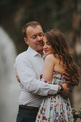 Young cute couple honeymoon standing near waterfall holding their hands on dating in a beautiful place italy near ocean and mountains, hug, smile and talk to each other
