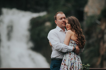 Young cute couple honeymoon standing near waterfall holding their hands on dating in a beautiful place italy near ocean and mountains, hug, smile and talk to each other
