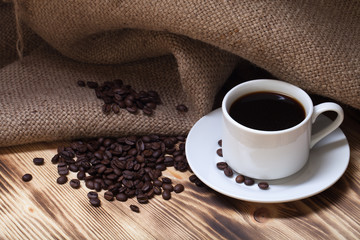Coffee beans and coffee in white cup on wooden table with burlap