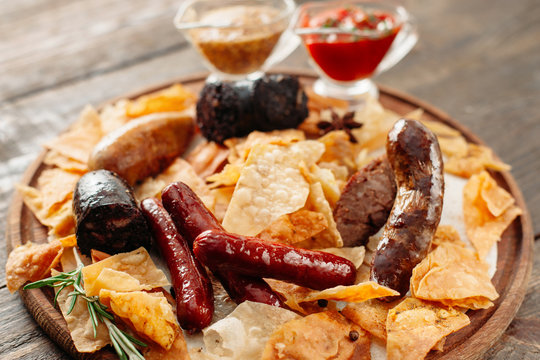 Beer snacks set on wooden plate. Closeup of wooden catering platter with assorted snacks for beer with two sauces. Different sausages and chips on wooden background. Focus on foreground