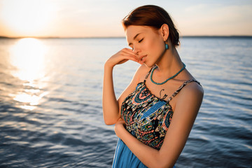 Fototapeta premium Young beautiful girl in blue dress posing at sea coast against blue sky at sunset