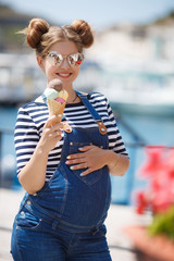 Beautiful pregnant brunette woman with trendy hairstyle,beautiful smile,dressed in blue denim overalls and striped t-shirt,mirrored sunglasses,posing on the promenade with a cone of colored ice cream