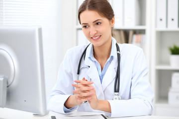 Young brunette female doctor sitting at the table and working at hospital office.  Health care, insurance and help concept. Physician ready to examine patient