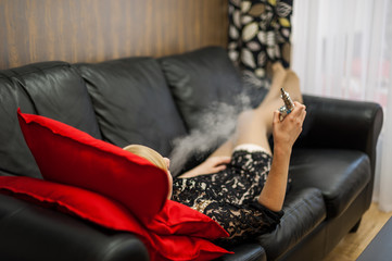 девушка на черном кожаном диване курит электронную сигарету . Girl on a black leather couch is smoking an electronic cigarette - obrazy, fototapety, plakaty