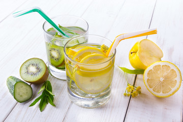 Fruit water in a glass of next to a lemon, kiwi, cucumber, apple, mint, linden.