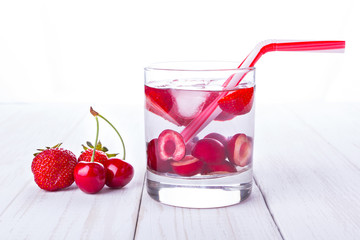 A glass of water with fruit, next to strawberries and cherries, tube on a wooden table