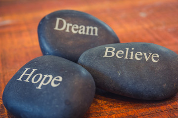 Black inspirational pebble stones with the words Dream, Believe and Hope on wooden background
