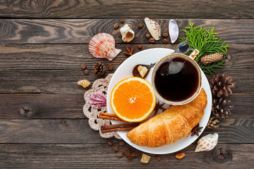 Continental breakfast - cup of hot coffee, croissant and orange. Tasty food on rustic wooden background with sea shells. Top view, place for text.