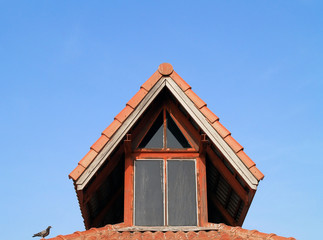 Fototapeta na wymiar outside of old house attic with glass window red wood frame and bird perched on orange roof tiles, close up small garret room with triangle pitched roof of vintage home and clear blue sky background