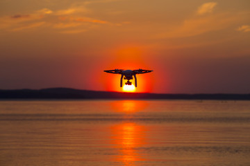 Fototapeta na wymiar Quad copter flying above water during a very red sunset. The copter is a silhouette against the sundown.