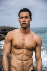 Young shirtless athletic man standing in water by ocean shore