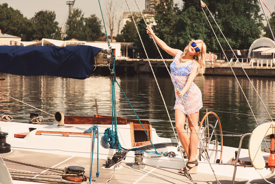 Young beautiful blond woman in white transparent dress and swimsuit standing on yacht at sunny day