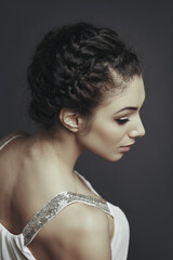 Side face portrait of beautiful female fashion model posing with braided hairdo, wearing white low cut blouse, looking down over grey background