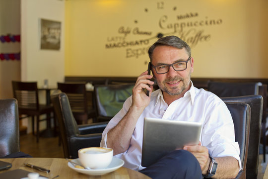 Portrait of smiling businessman with digital tablet sitting in a cafe telephoning with smartphone