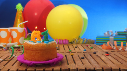 Birthday cake with candles on rustic wooden table with background of colorful balloons, gifts,...