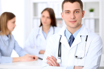 Fototapeta premium Friendly male doctor on the background with patient and his doctor in hospital. High level and quality medical service concept.